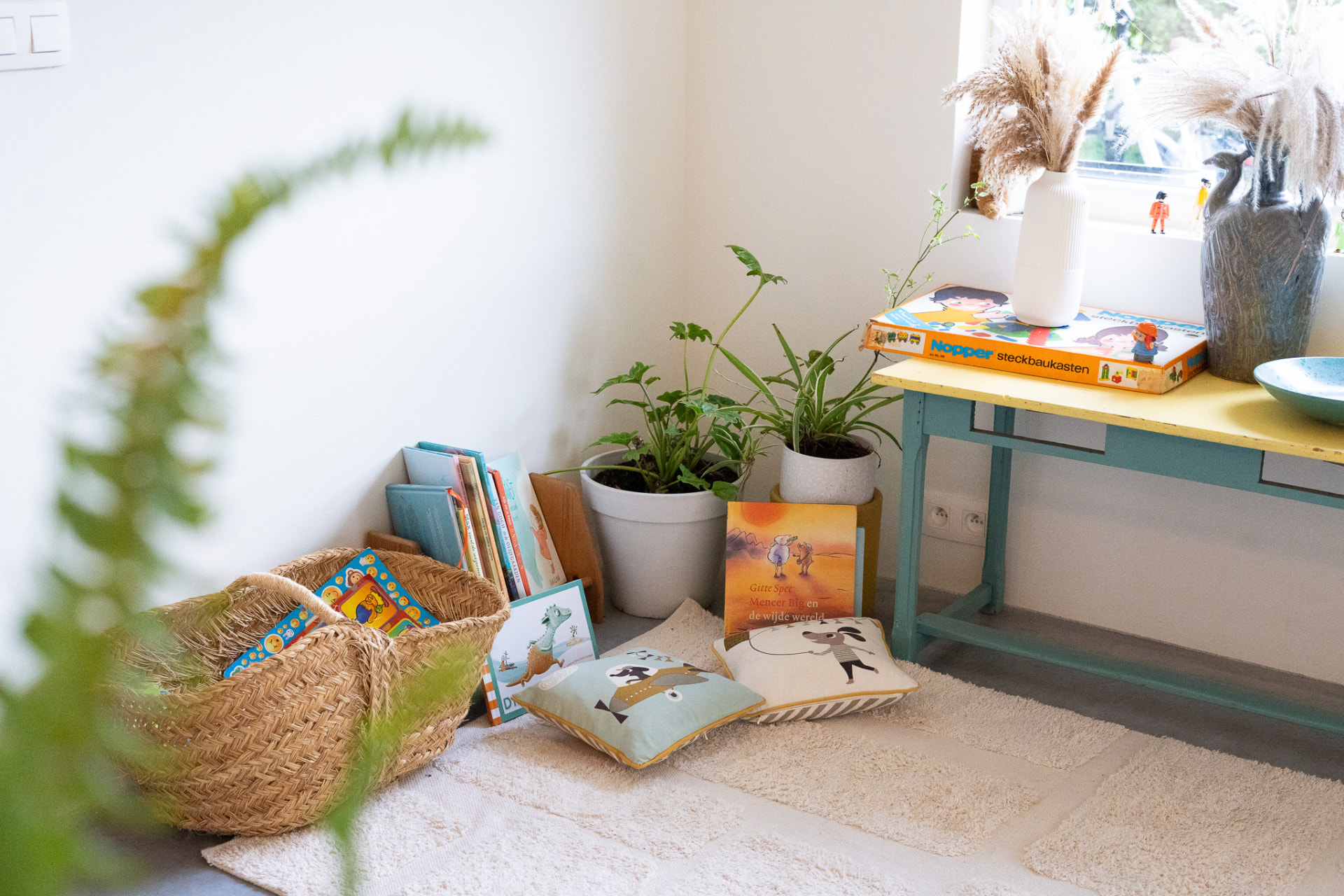 Lichte speelhoek met een rieten mand vol prentenboeken, twee kussens op een zacht kleed, kamerplanten en een lichtblauw kinderbureautje met geel blad